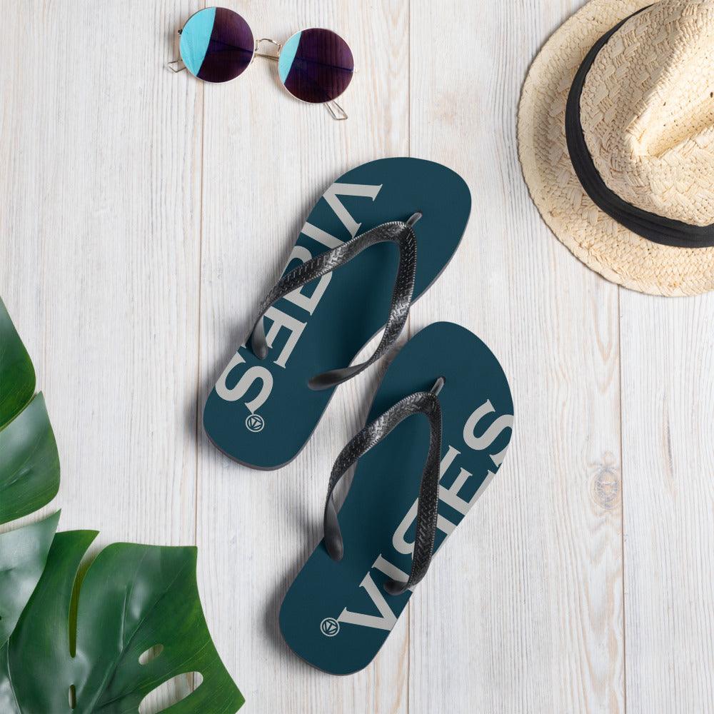 Blaue VIBES Flip-Flops auf hellem Holz mit Sonnenbrille, Strohhut und Monstera-Blatt