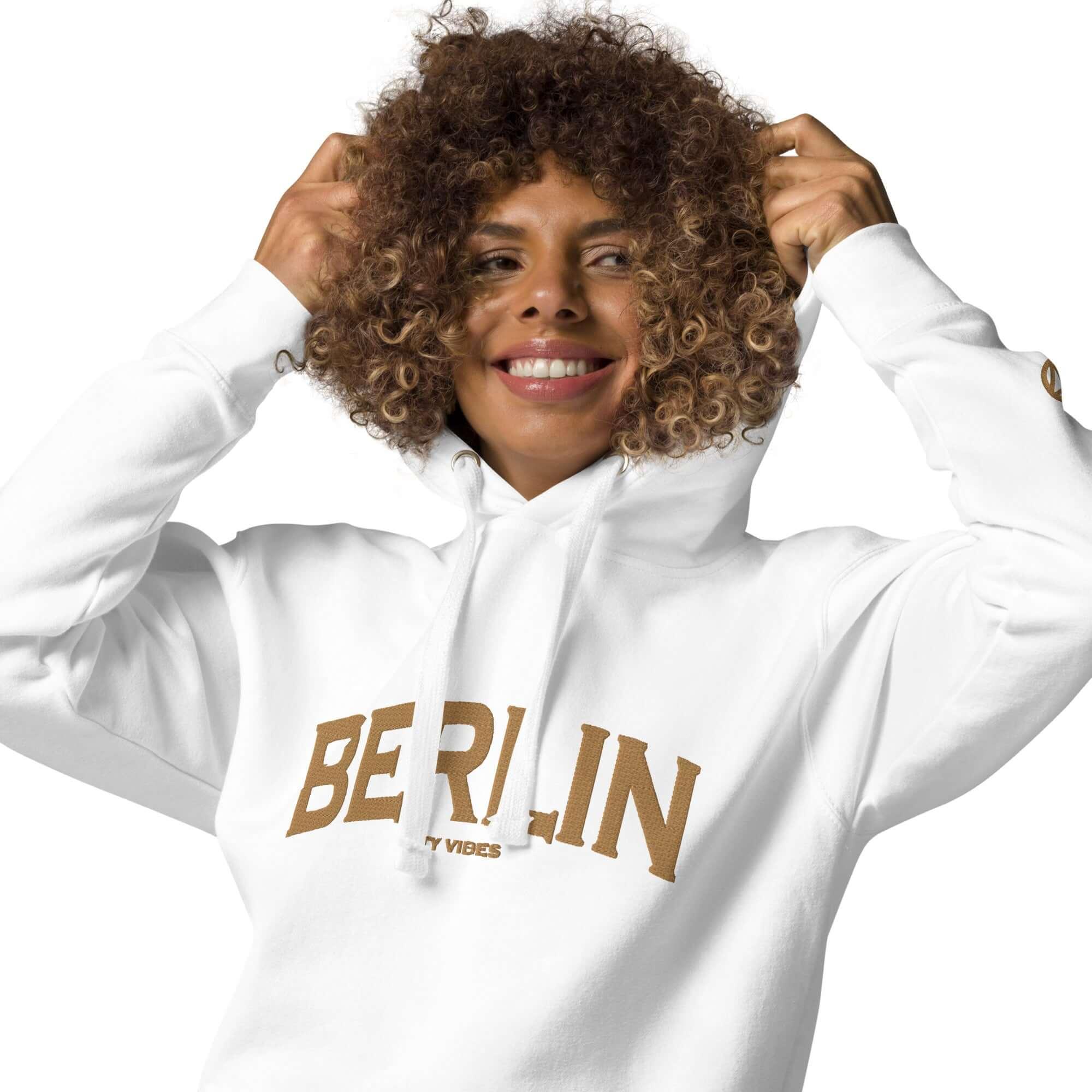 Frau mit lockigem Haar trägt weißen CREWILD Hoodie mit goldenem Berlin-Schriftzug.