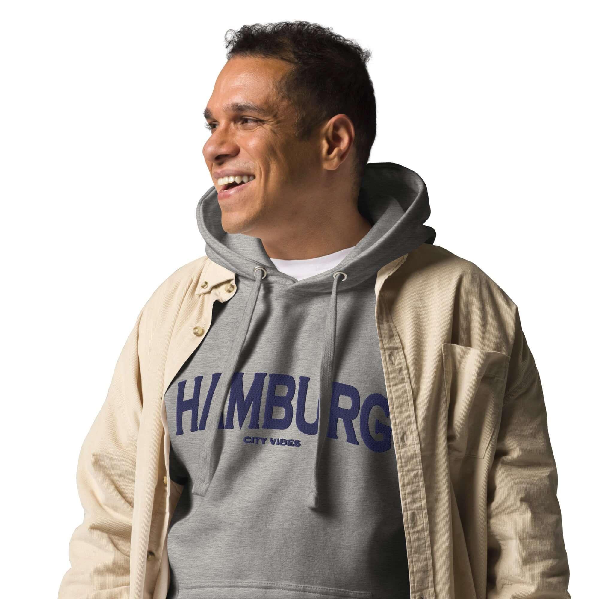 Lachender Mann im grauen CREWILD Hoodie mit blauem Hamburg-Print und beiger Jacke.