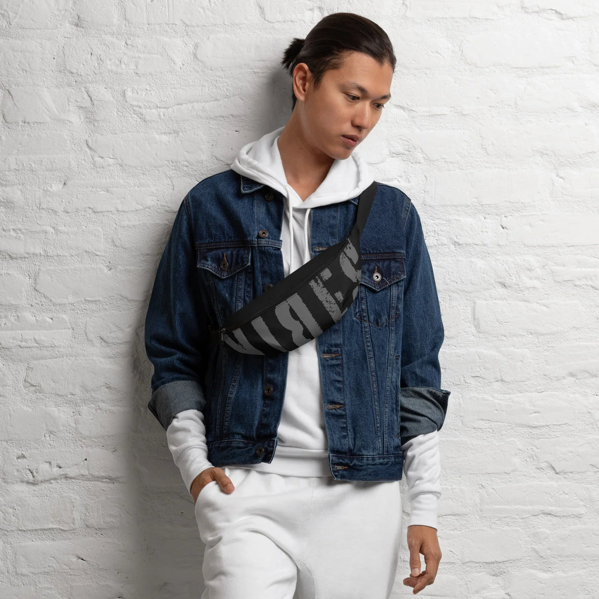 Mann in weißem Hoodie, Jeansjacke und schwarzer Bauchtasche vor weißer Ziegelwand