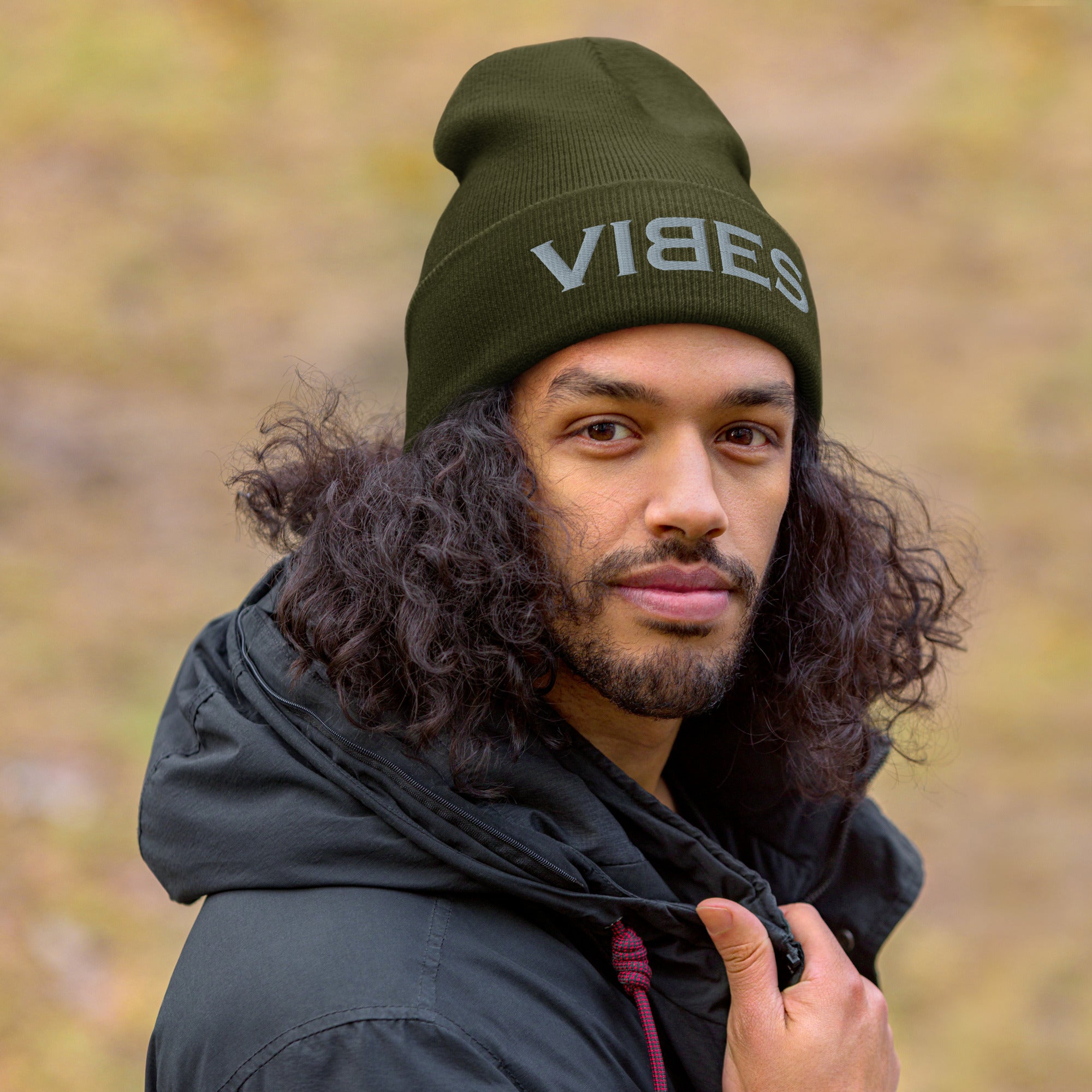 Mann mit lockigen Haaren trägt eine olivgrüne CREWILD Beanie Mütze mit grauem VIBES-Stick.