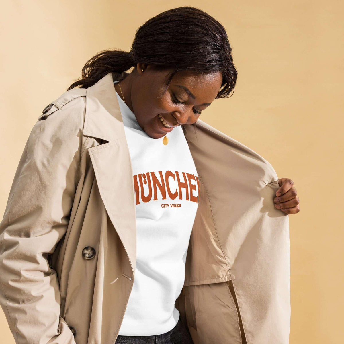 Frau mit beigem Trenchcoat und weißem München City Vibes T-Shirt vor beigem Hintergrund