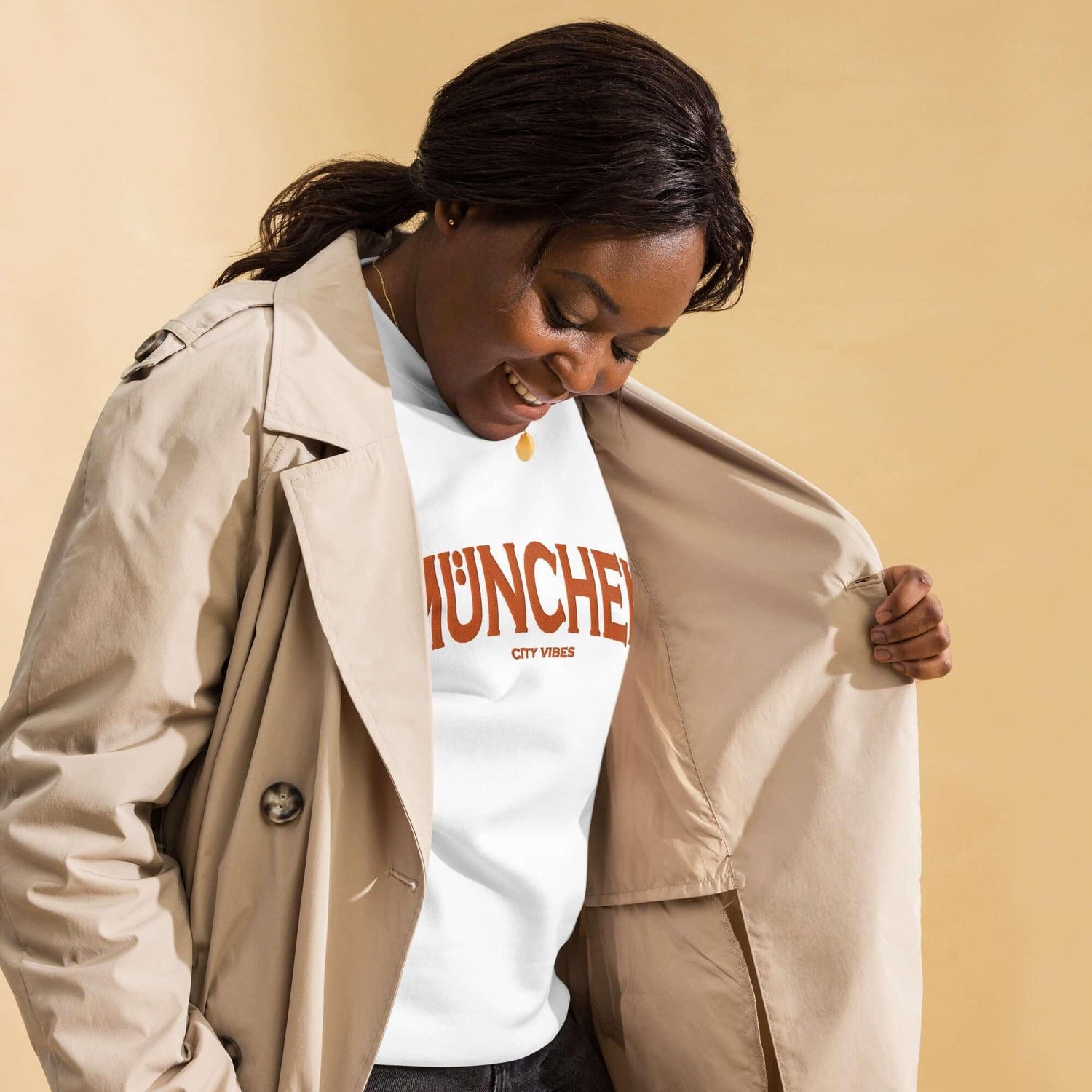 Frau mit beigem Trenchcoat und weißem München City Vibes T-Shirt vor beigem Hintergrund