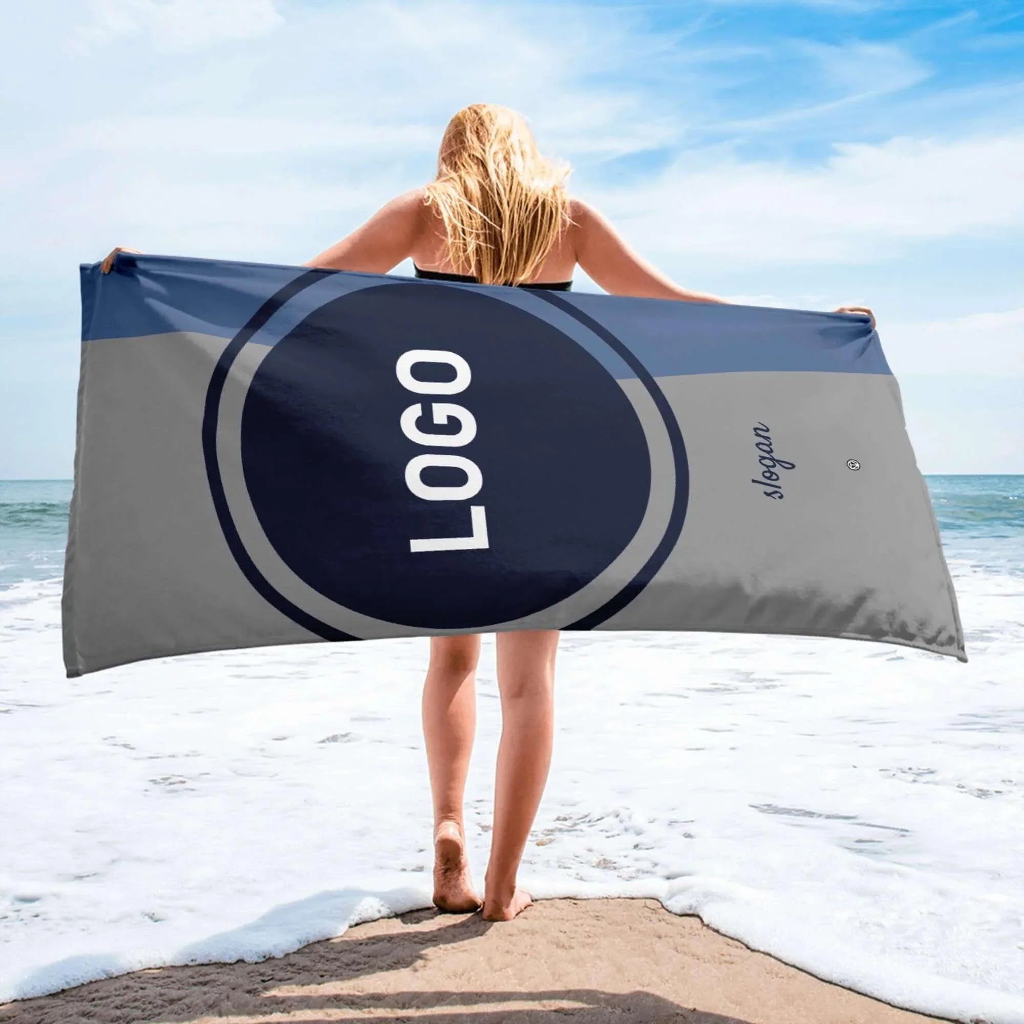 Frau am Strand hält graues und blaues Werbehandtuch mit Logo-Aufdruck am Meer