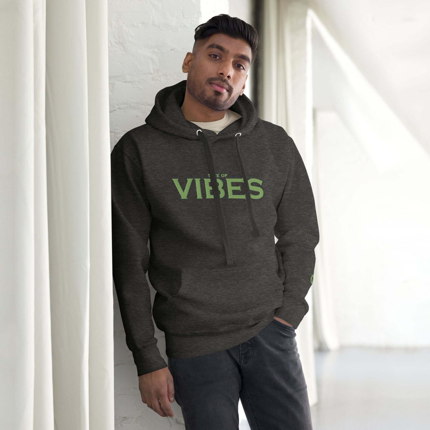 Mann trägt grauen Unisex-Hoodie mit grünem Vibes-Print, steht vor weißer Wand
