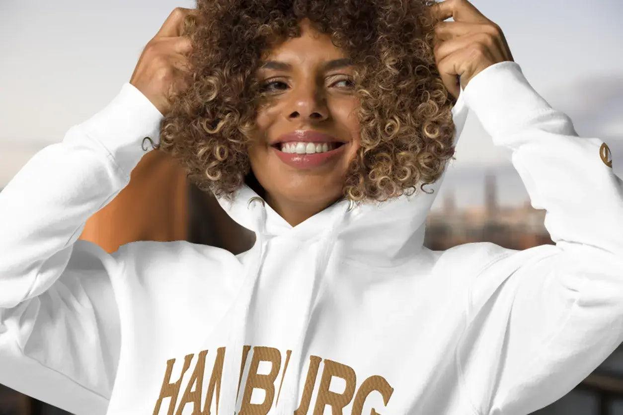 Frau mit lockigen Haaren trägt weißen Hamburg Hoodie und lächelt, urbaner Hintergrund.