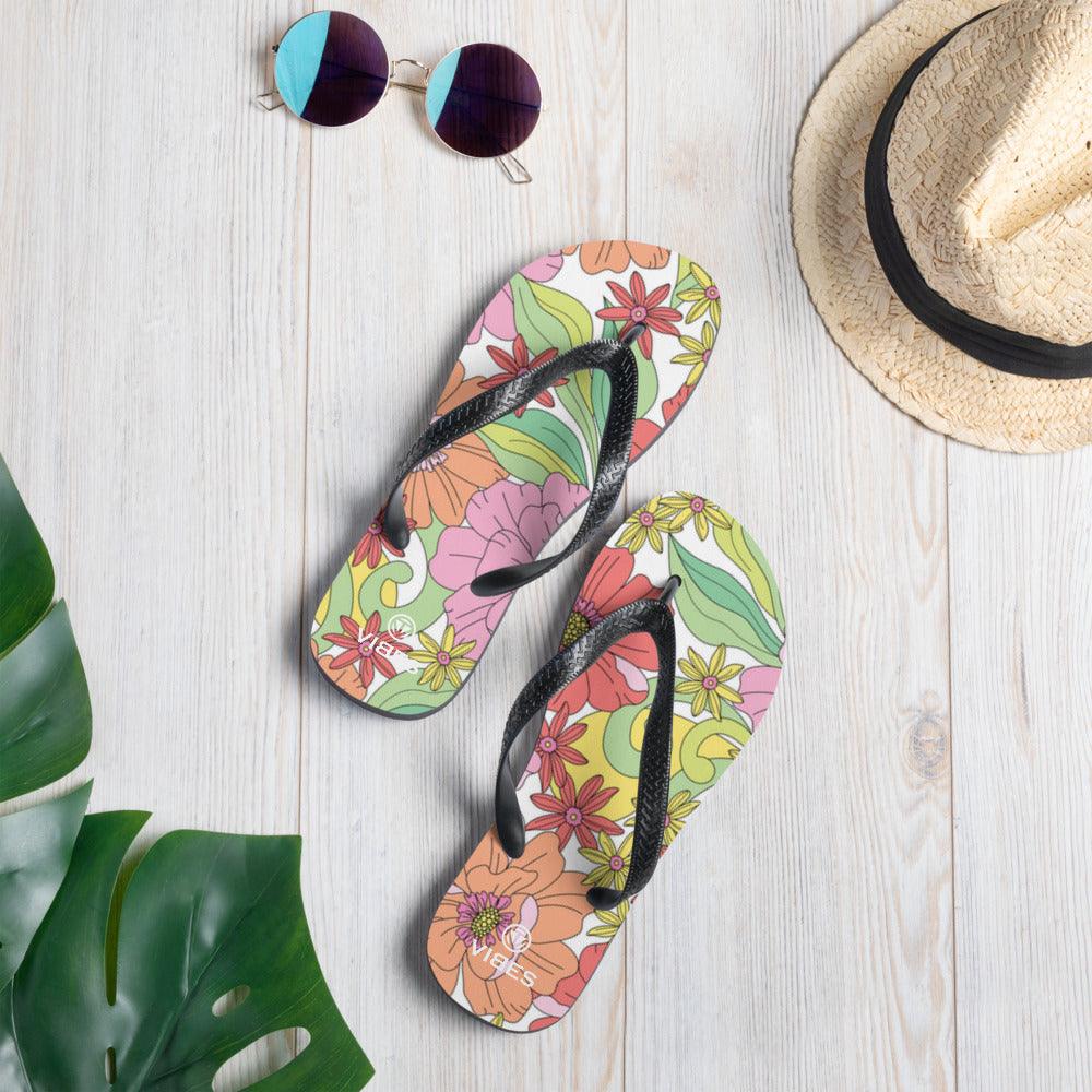 Bunte VIBES Flip-Flops mit Blumenmuster auf hellem Holz, Sonnenbrille und Strohhut daneben