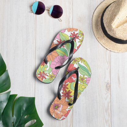 Bunte VIBES Flip-Flops mit Blumenmuster auf hellem Holz, Sonnenbrille und Strohhut daneben