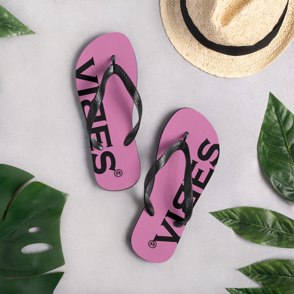 Pink Flip Flops mit schwarzem VIBES Schriftzug auf grauem Hintergrund, Strohhut und Blättern