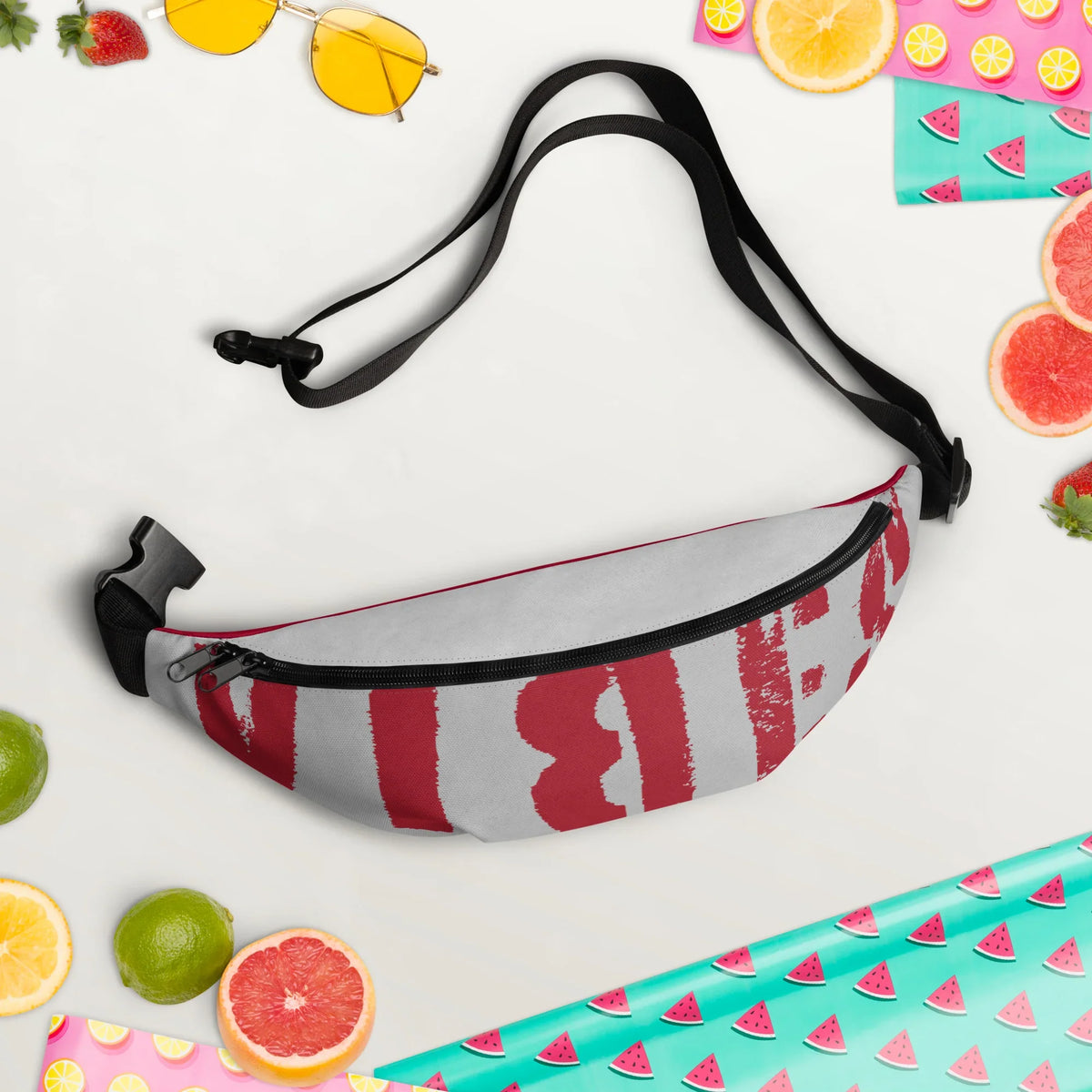 Graue Gürteltasche mit rotem Grafikprint, umgeben von sommerlichen Accessoires und Früchten