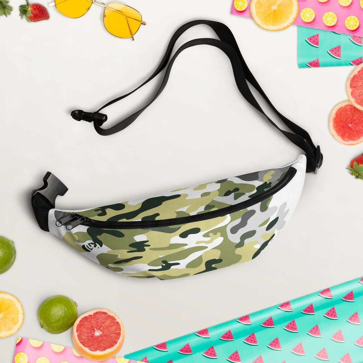 Camouflage-gemusterte Gürteltasche auf weißem Hintergrund mit sommerlichen Früchten und Accessoires
