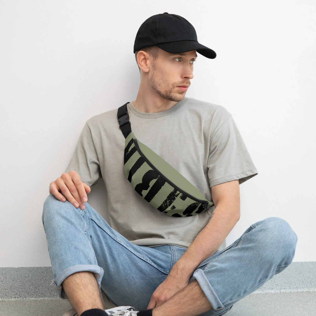 Mann mit schwarzer Cap, grauem T-Shirt und einer olivgrünen Gürteltasche mit schwarzem Print.