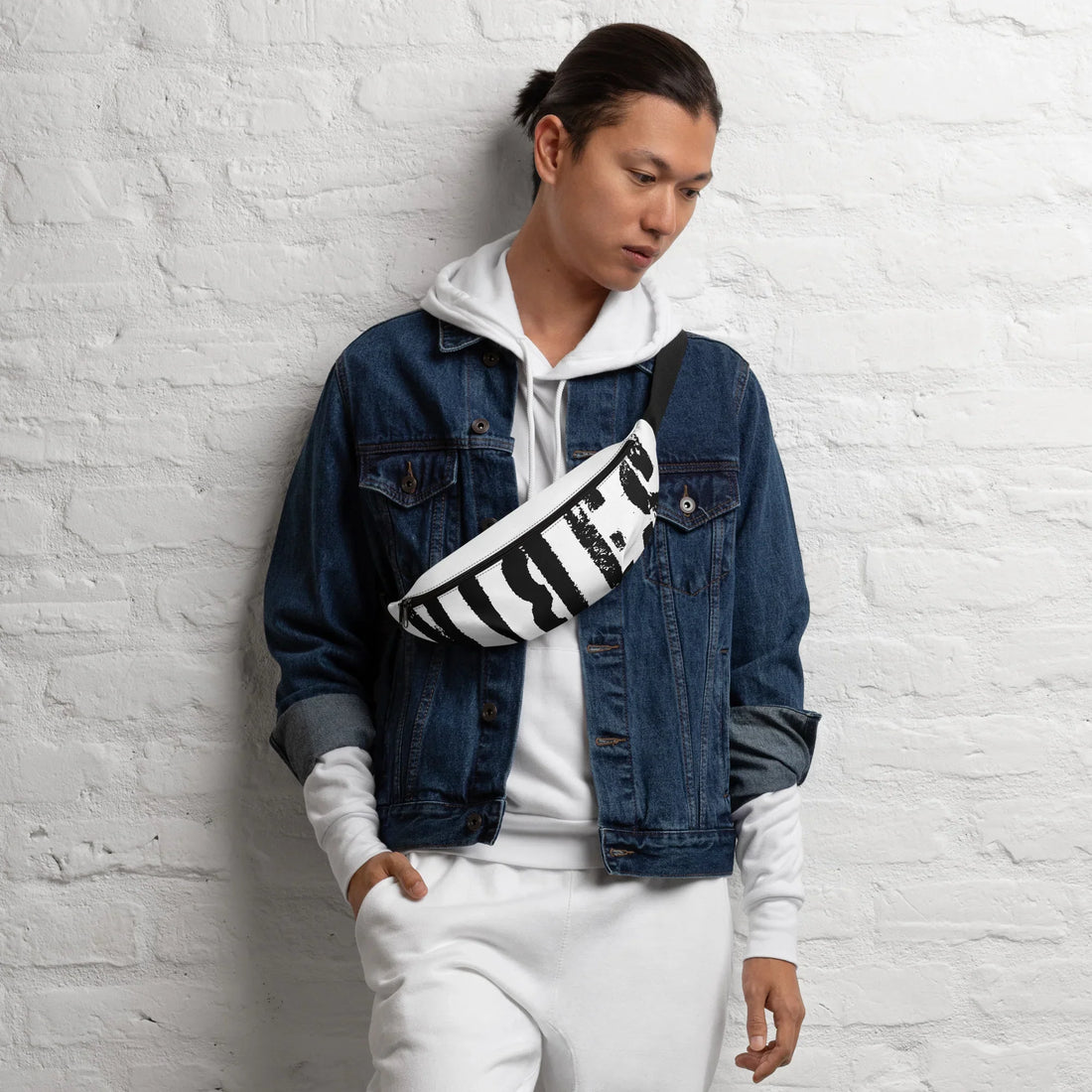 Mann mit weißem Hoodie, Jeansjacke und einer schwarz-weißen Gürteltasche vor weißer Ziegelwand