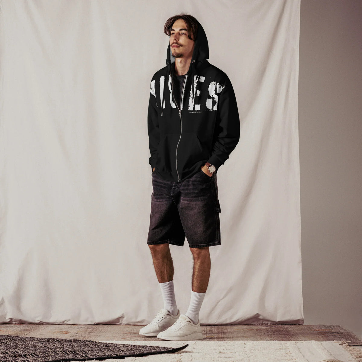 Junger Mann in schwarzem Hoodie mit weißem Print, dunklen Shorts und weißen Sneakern.