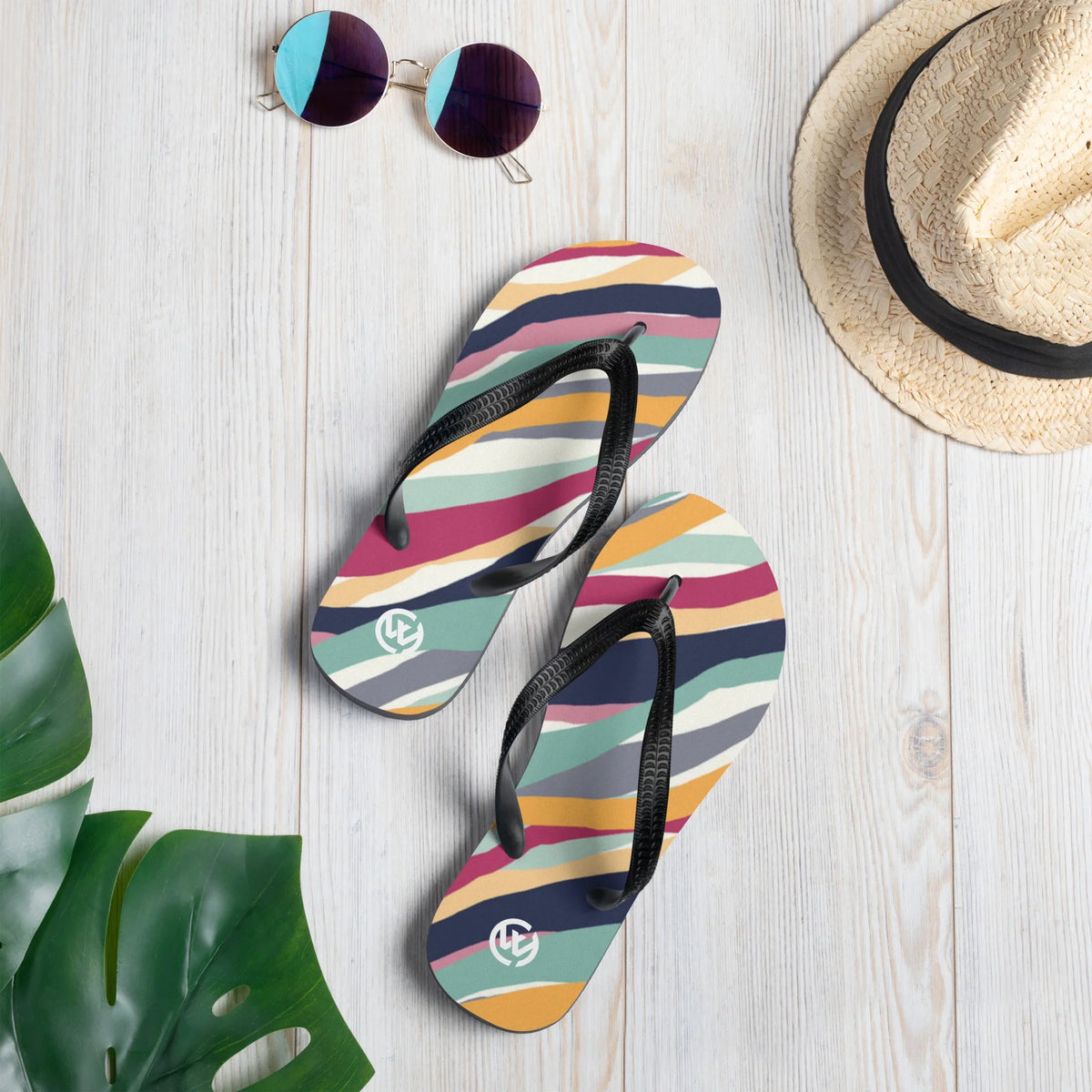 Bunte CREWILD Flip-Flops mit Streifenmuster neben Sonnenbrille und Strohhut auf hellem Holz.