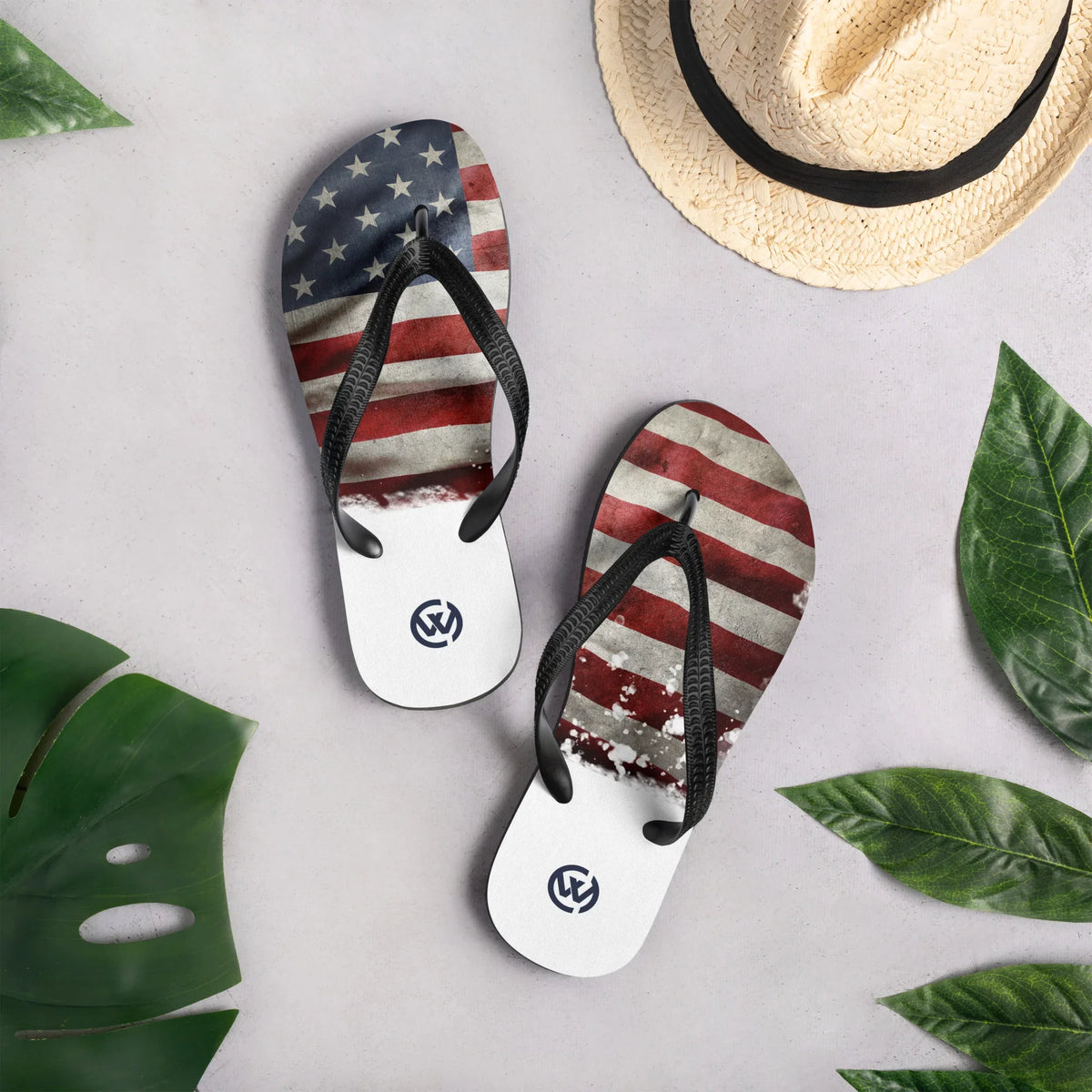 CREWILD Flip-Flops mit US-Flaggen-Design auf grauem Hintergrund mit Strohhut und Blättern
