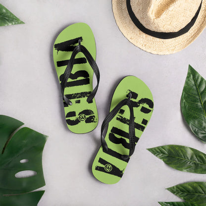 Grüne CREWILD Flip-Flops mit schwarzem VIBES-Schriftzug neben einem Strohhut und Blättern.