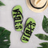 Grüne CREWILD Flip-Flops mit schwarzem VIBES-Schriftzug neben einem Strohhut und Blättern.