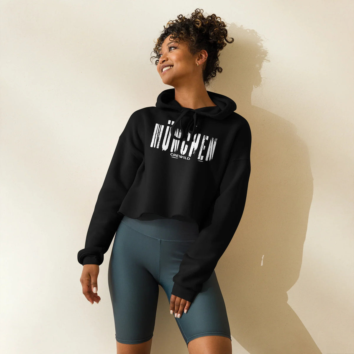 Frau trägt schwarzen CREWILD Crop-Hoodie mit weißem München-Schriftzug und graue Biker-Shorts.