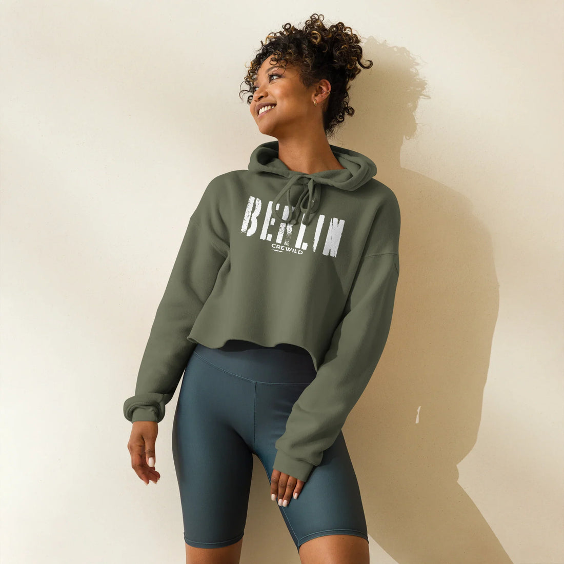 Frau trägt olivgrünen CREWILD Crop-Hoodie mit weißem Berlin-Schriftzug und dunkelblaue Shorts.