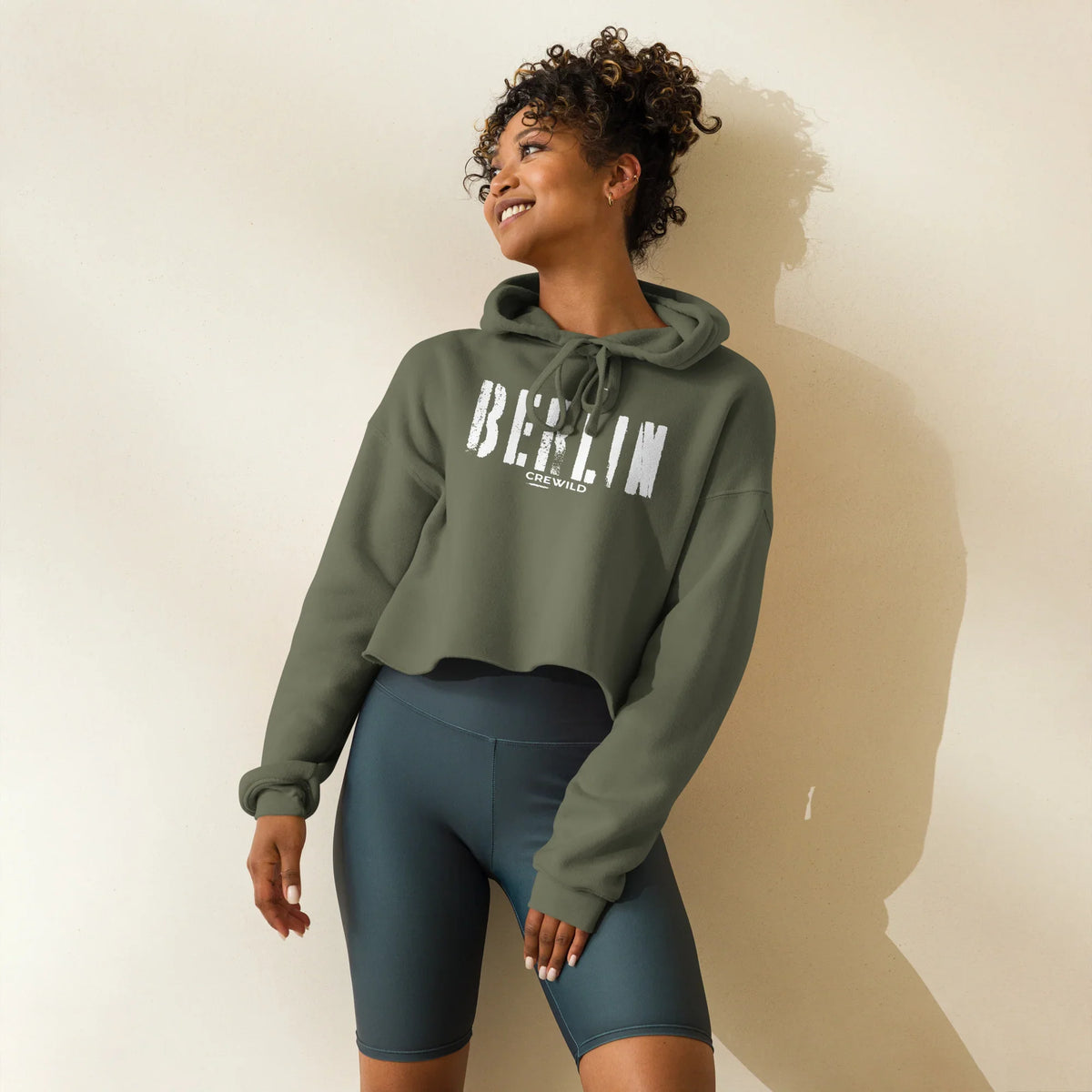 Frau trägt olivgrünen CREWILD Crop-Hoodie mit weißem Berlin-Schriftzug und dunkelblaue Shorts.