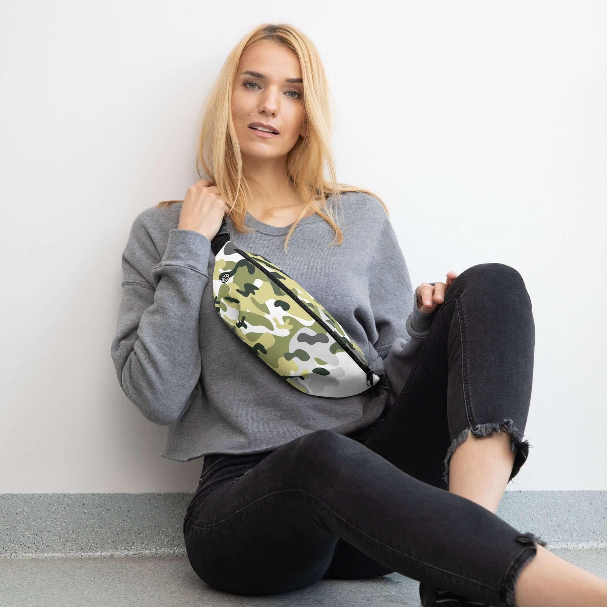 Frau mit grauem Pullover und Camouflage-Bauchtasche sitzt entspannt an weißer Wand