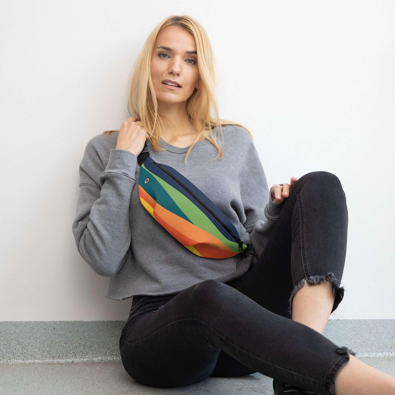 Frau mit bunter Bauchtasche, grauem Pullover und schwarzer Jeans sitzt vor weißer Wand