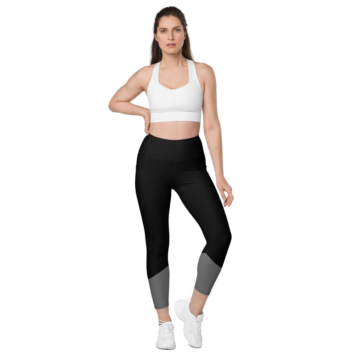 Frau trägt schwarze Leggings mit Taschen, weißem Sport-BH und weißen Turnschuhen vor weißem Hintergrund