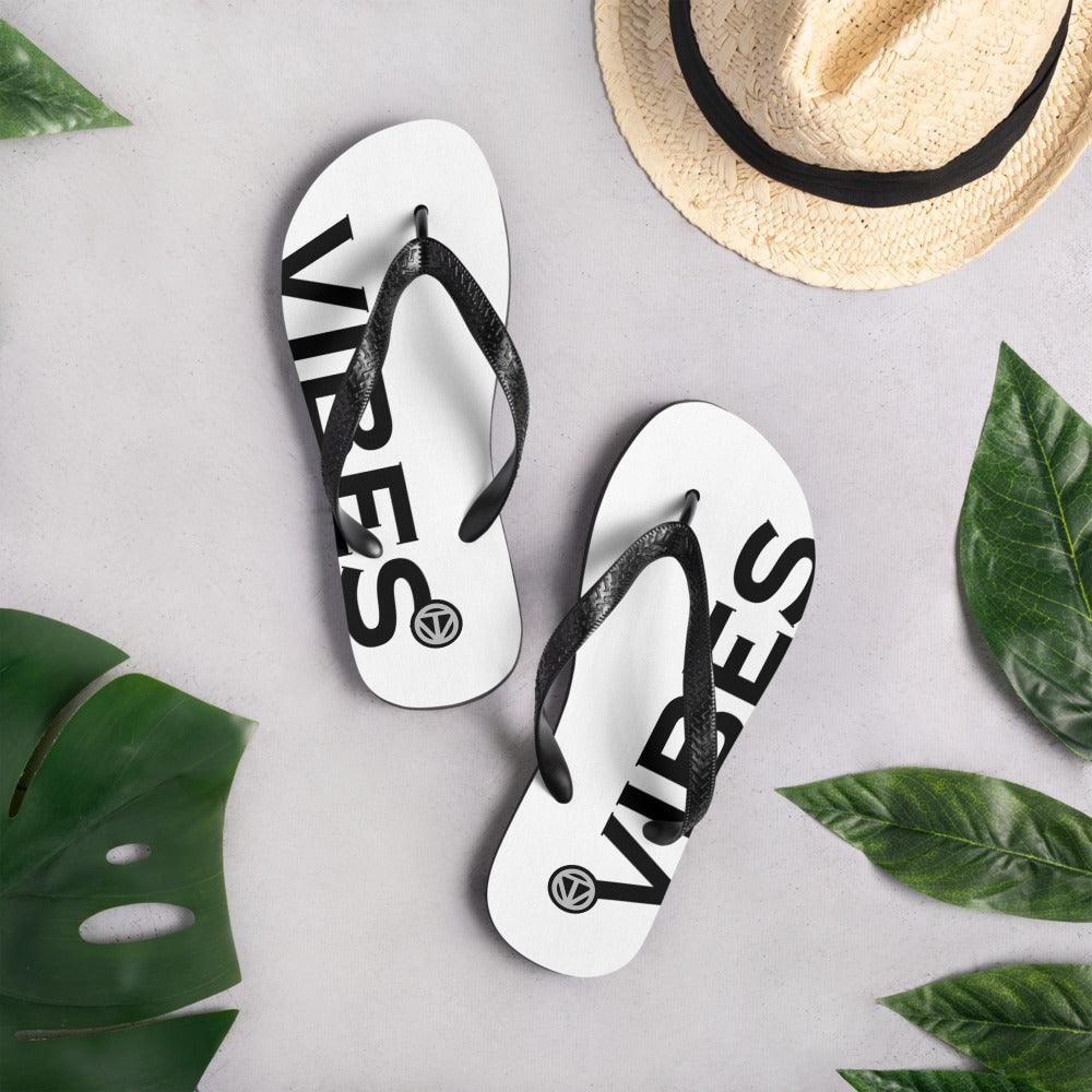 Weiße Flip Flops mit schwarzem Vibes-Print, Strohhut und grünen Blättern auf hellem Untergrund