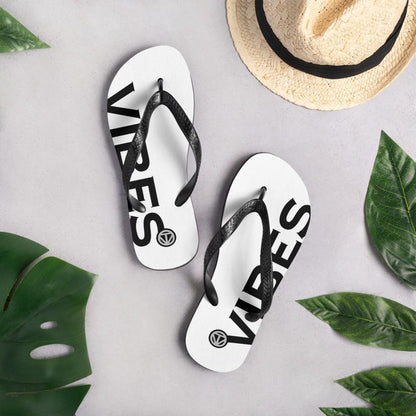 Weiße Flip Flops mit schwarzem Vibes-Print, Strohhut und grünen Blättern auf hellem Untergrund
