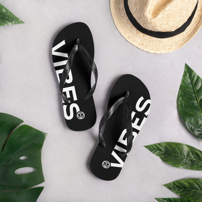 Schwarze Flip-Flops mit weißem Vibes-Print, Sommerhut und tropischen Blättern auf hellem Untergrund