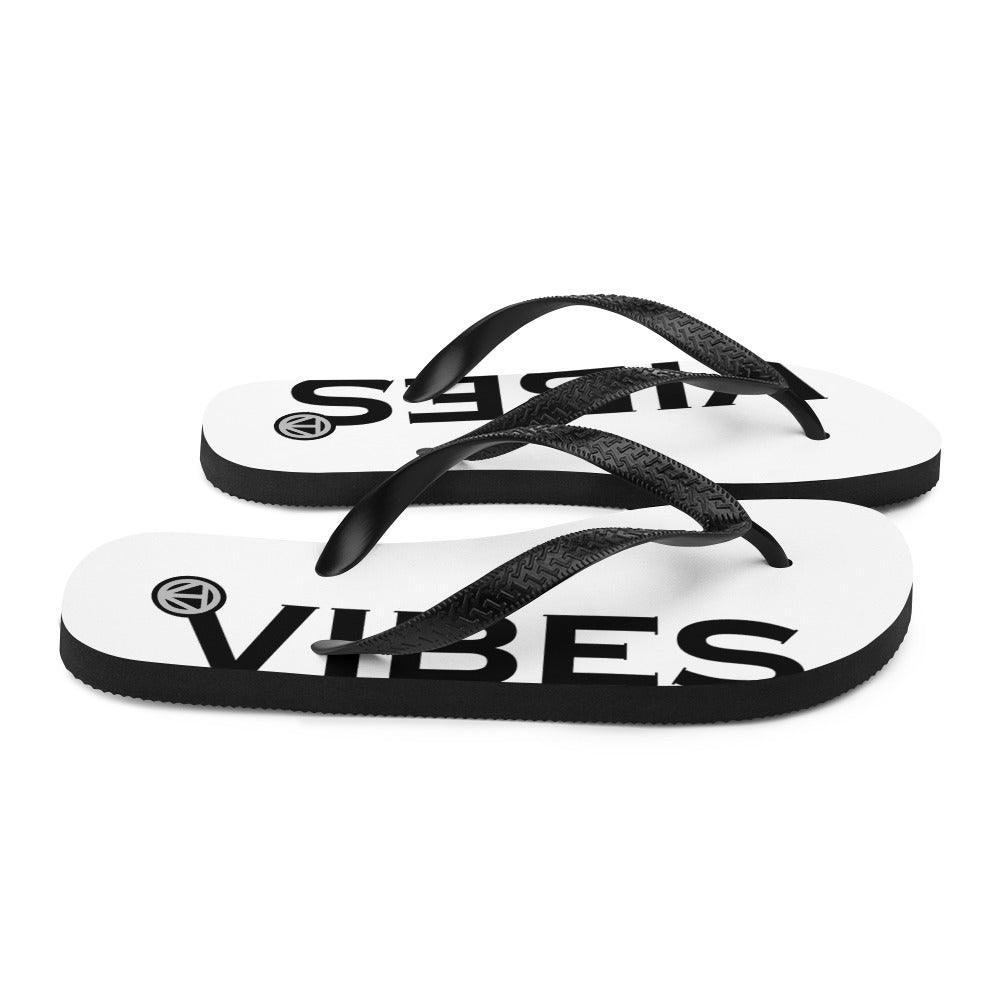 Weiße Flip Flops mit schwarzem VIBES Print und schwarzen Riemen auf weißem Hintergrund